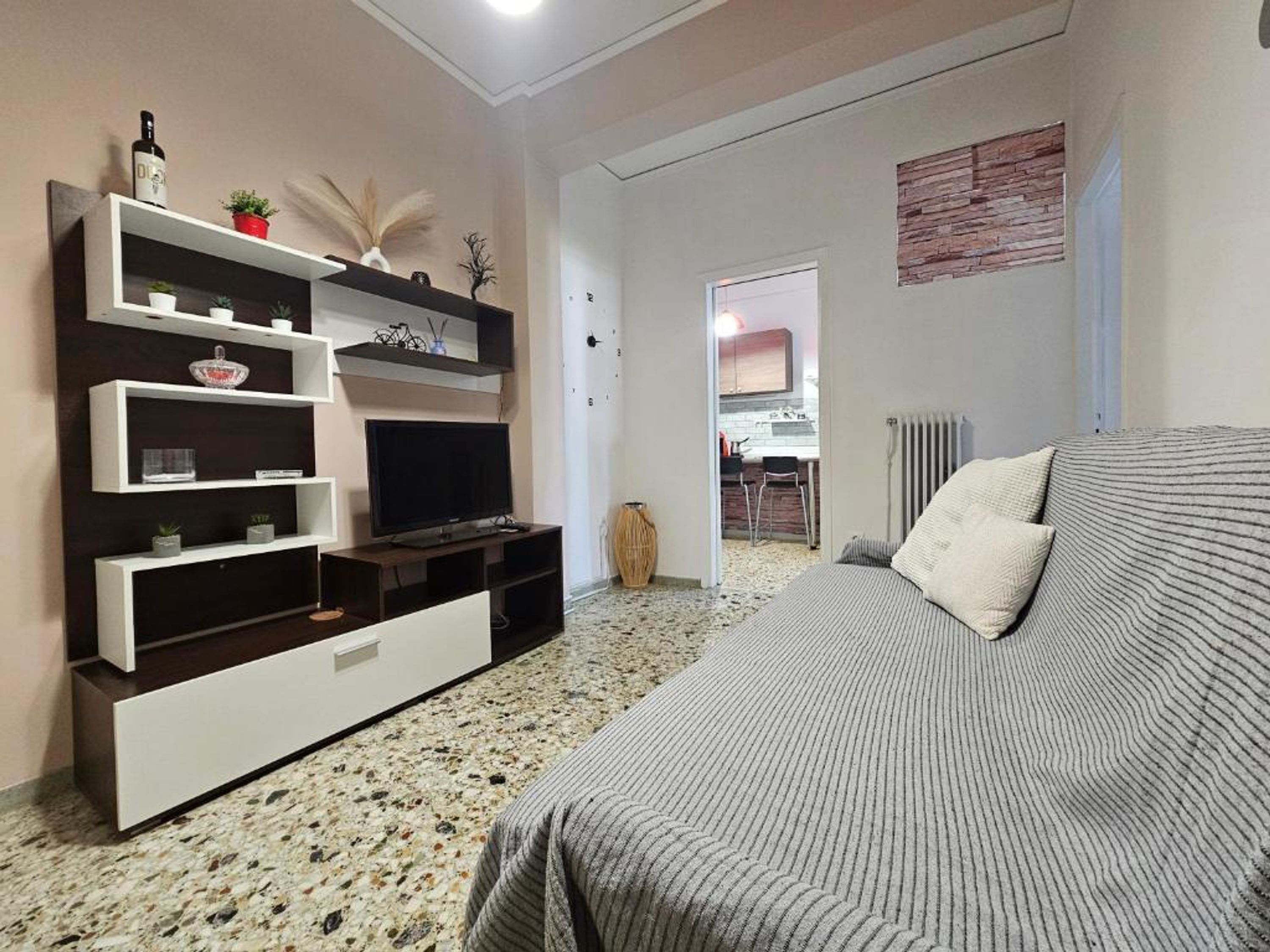 Een slaapkamer met een bed, een televisie op een standaard, een keuken op de achtergrond, en een gang naar een andere kamer.