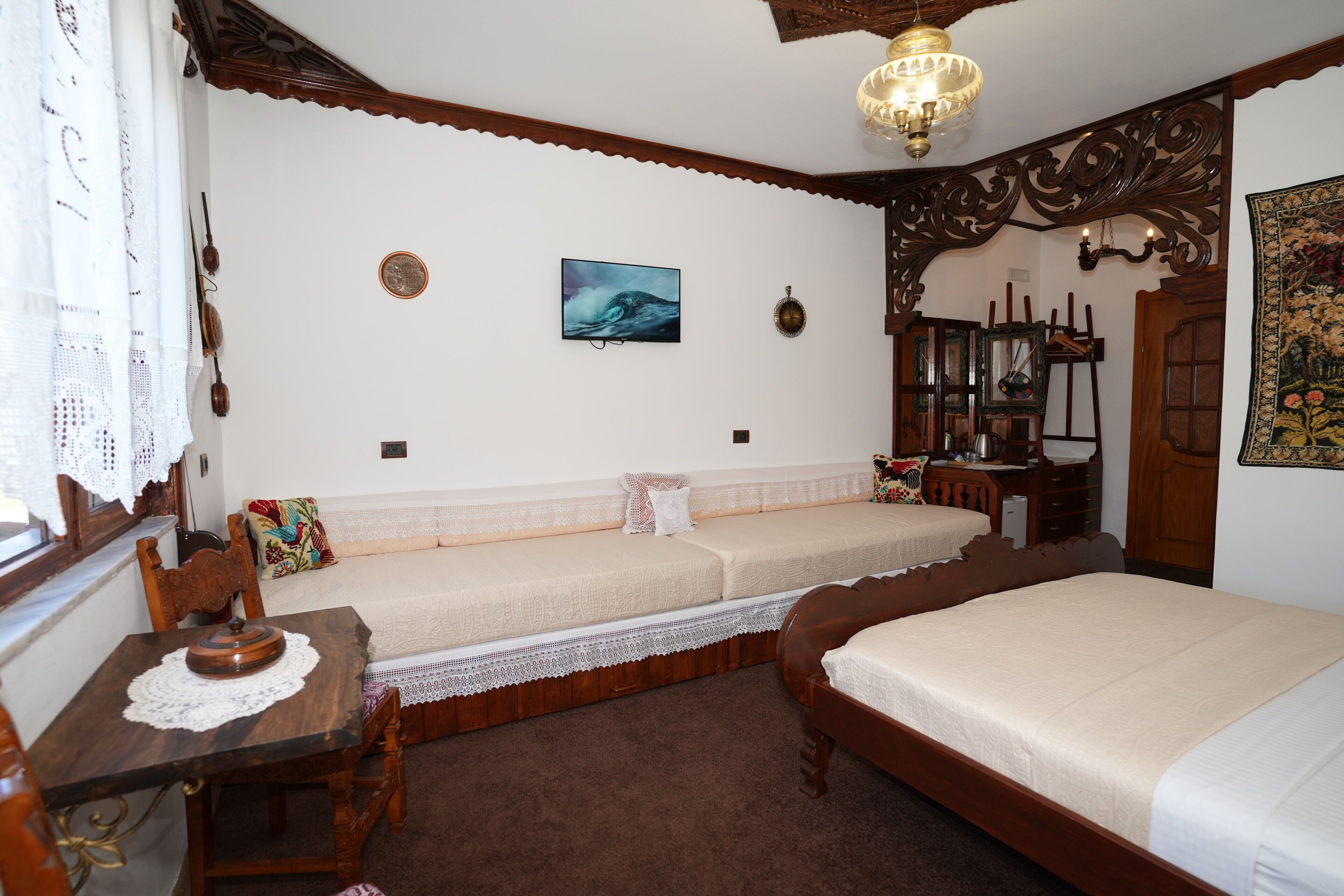 Una habitación con una cama de madera, una mesa de madera con mantel de encaje, un banco con cojines, un cuadro colgado en la pared y una lámpara tradicional.