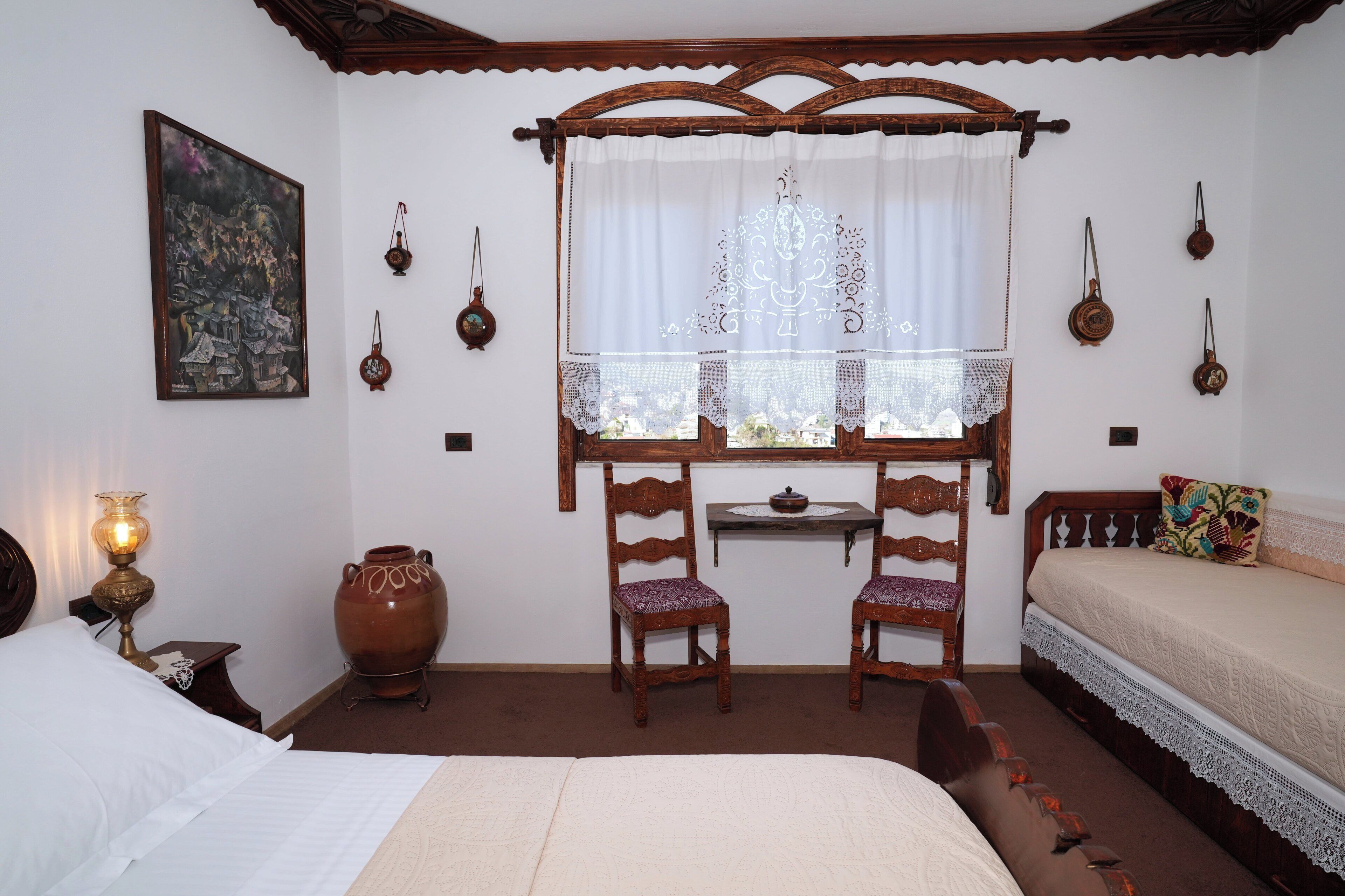 Une chambre traditionnelle comprenant un lit, deux chaises, une table, une fenêtre avec des rideaux et des lampes fixées au mur.