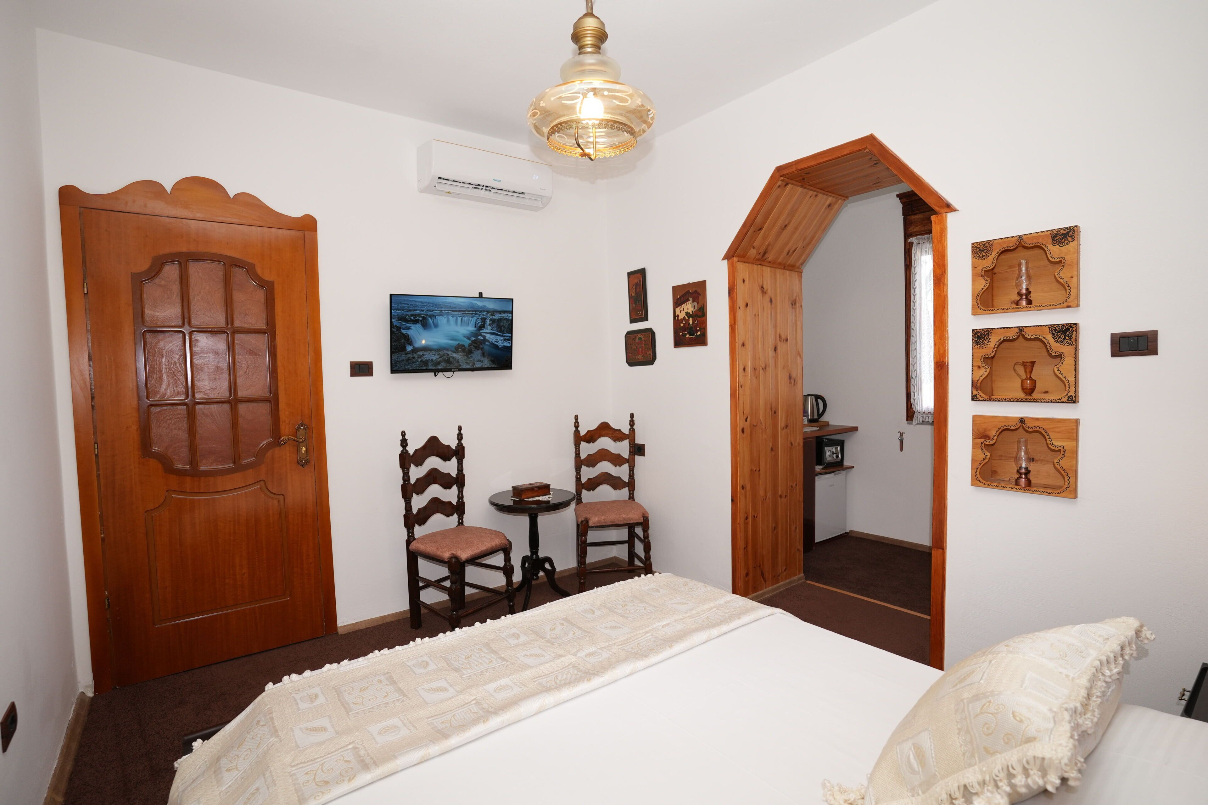 A bedroom with a wooden door, two chairs, a bed with a patterned bedspread, a ceiling light, and a wall-mounted TV.