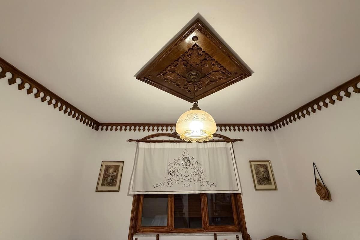 Ein Zimmer mit Holzboden, einer Zierleuchte, einem Fenster mit Vorhang und gerahmten Bildern an den Wänden.