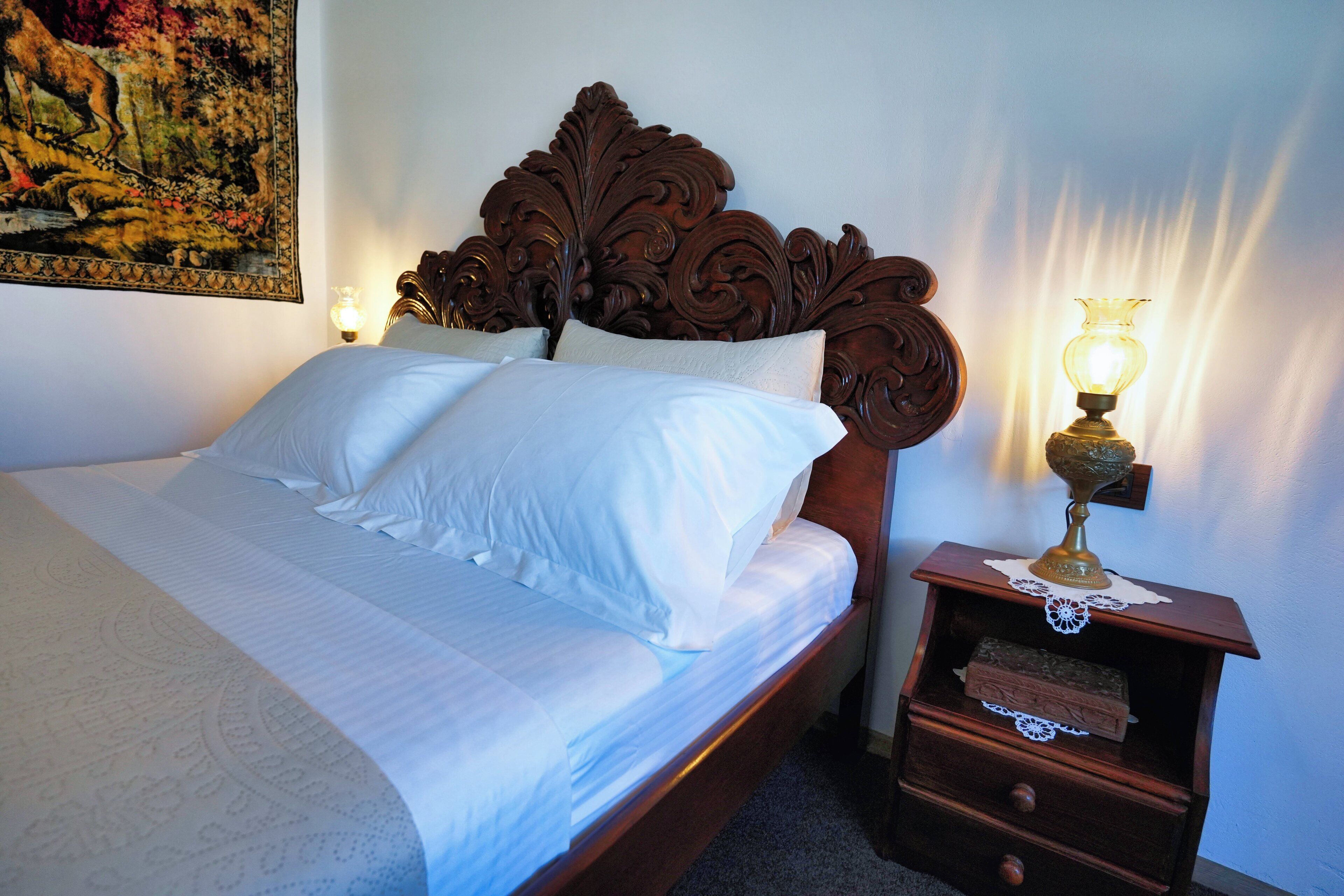 A neatly made bed with white linens, a wooden headboard, a bedside table with a lamp, and a wall-mounted tapestry.