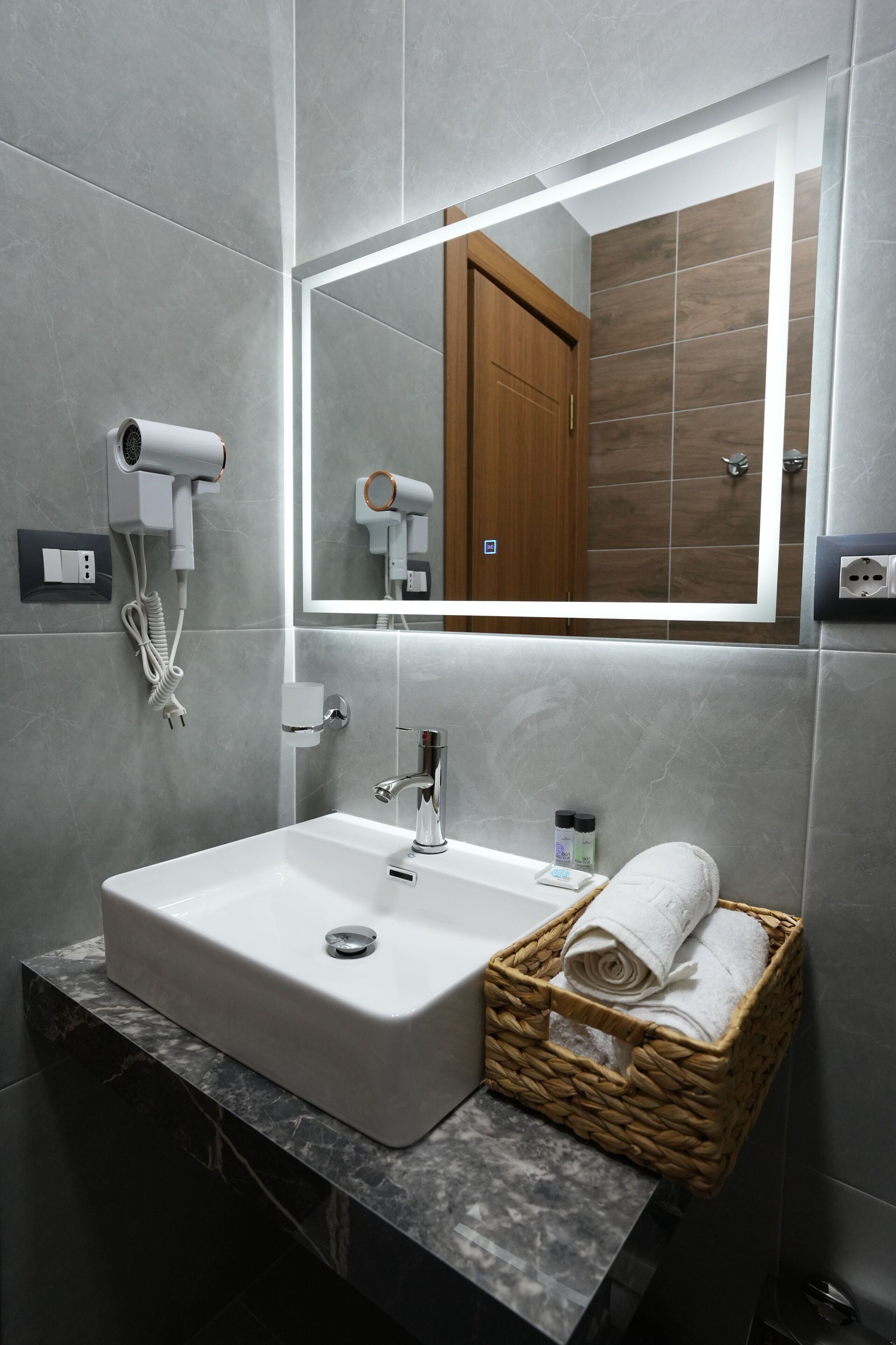 A bathroom with a white sink, a mirror, a hairdryer, a towel basket, and a wooden door.