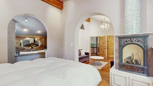 Een slaapkamer met een groot bed, een kaptafel met spiegel, een open haard en een gebogen deuropening naar een andere kamer.