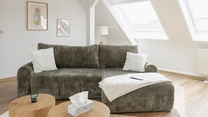 Un salon moderne avec un canapé vert, une table basse ronde en bois et un pouf blanc. Deux œuvres d’art encadrées ornent le mur, et une lampe sur pied est placée dans un coin.