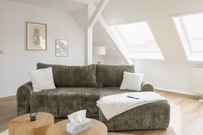 Un salon moderne avec un canapé vert, une table basse ronde en bois et un pouf blanc. Deux œuvres d’art encadrées ornent le mur, et une lampe sur pied est placée dans un coin.