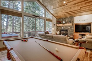 Ein Billardtisch mit Spielzeug und Stöcke, eine Couch, ein Fernseher und ein Kamin aus Stein in einem Raum mit großen Fenstern und Blick auf Bäume.