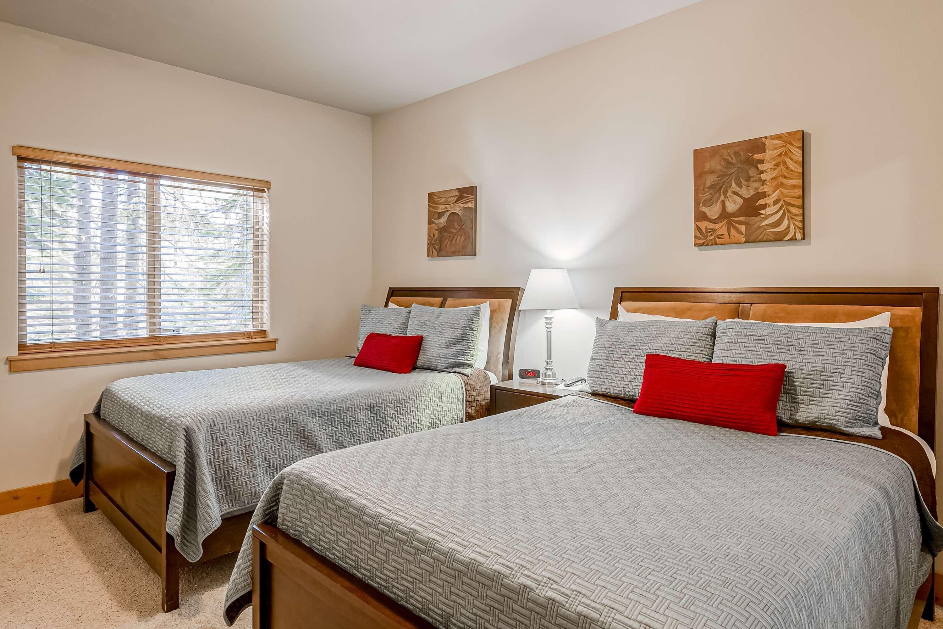 A bedroom with two beds, a window with blinds, a nightstand with a lamp, and wall art.