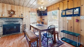 Un interior de madera con chimenea, una mesa de comedor con sillas, un banco, un televisor montado en la pared y un ventanal con vista a árboles.
