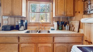 Cocina con armarios de madera, un lavamanos doble, una cafetera y un ventanal con vistas a árboles.