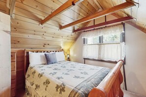Ein Schlafzimmer mit Holzboden, einem Bett mit floralem Überwurf, einem Nachttisch, einem Fenster mit Vorhängen und einem Holzkopfende.