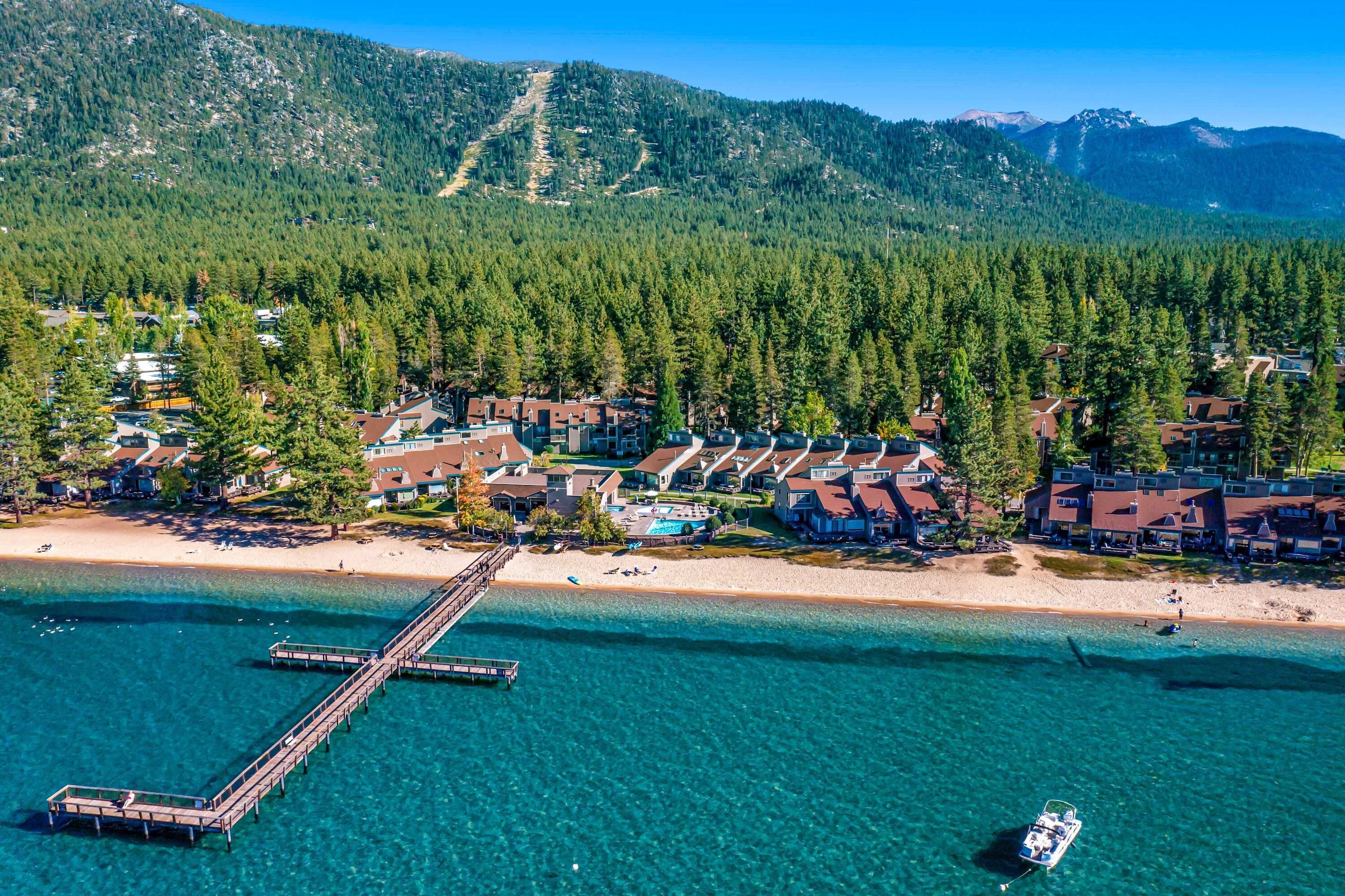 An aerial view of a coastal resort with a pier, clear turquoise waters, and a backdrop of dense forested mountains.