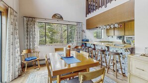 A dining area with a wooden table and chairs, a kitchen with a stone backsplash, and a view of trees through the window. - Spacious home with incredible views, full kitchen, shared  pool & tennis courts (Incline Village)