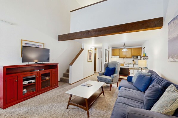 A living room with a red entertainment unit, a grey sofa, a white coffee table, and a kitchen in the background. - Lakefront retreat with shared pool, hot tub, & sauna - near shopping & skiing (South Lake Tahoe)