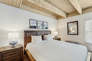 Une chambre à coucher avec une tête de lit en bois, un grand lit, deux tables de chevet avec des lampes, un banc, un miroir et des œuvres d’art encadrées accrochées au mur.