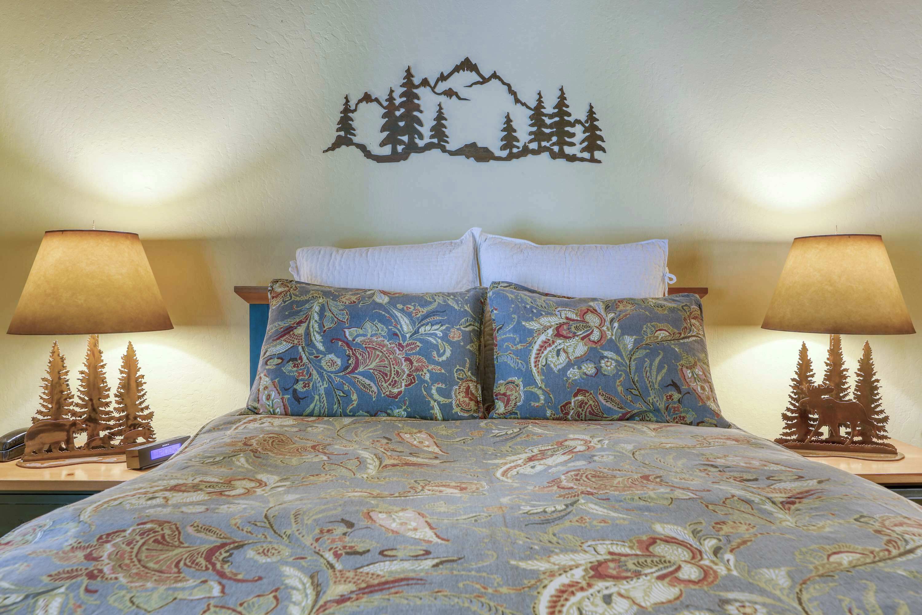 Un lit avec une literie à motifs, deux lampes de chevet décorées de motifs d’arbres et un décor mural représentant une montagne et des arbres.