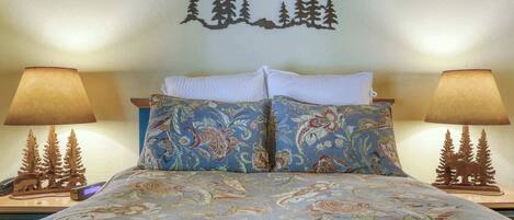 Un lit avec une literie à motifs, deux lampes de chevet décorées de motifs d’arbres et un décor mural représentant une montagne et des arbres.