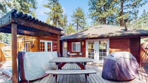 Een houten terras met een picknicktafel en overdekte barbecue, omringd door een houten chalet met een overdekt terras en uitzicht op hoge bomen.