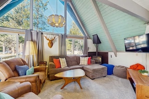 Ein geräumiges Wohnzimmer mit großem Fenster, Deckenventilator, Wand-Fernseher und Couchtisch aus Holz.