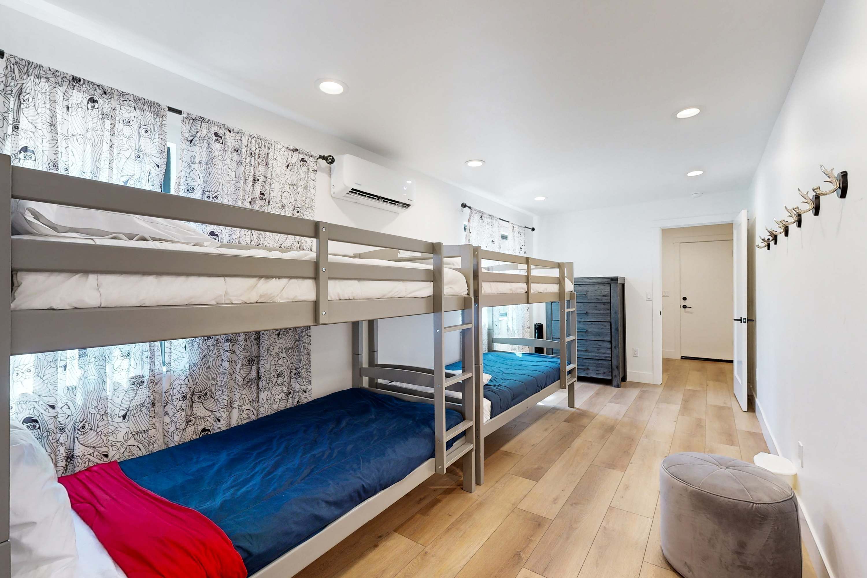 A bunk bed room with a patterned curtain, a grey ottoman, and a door in the background.