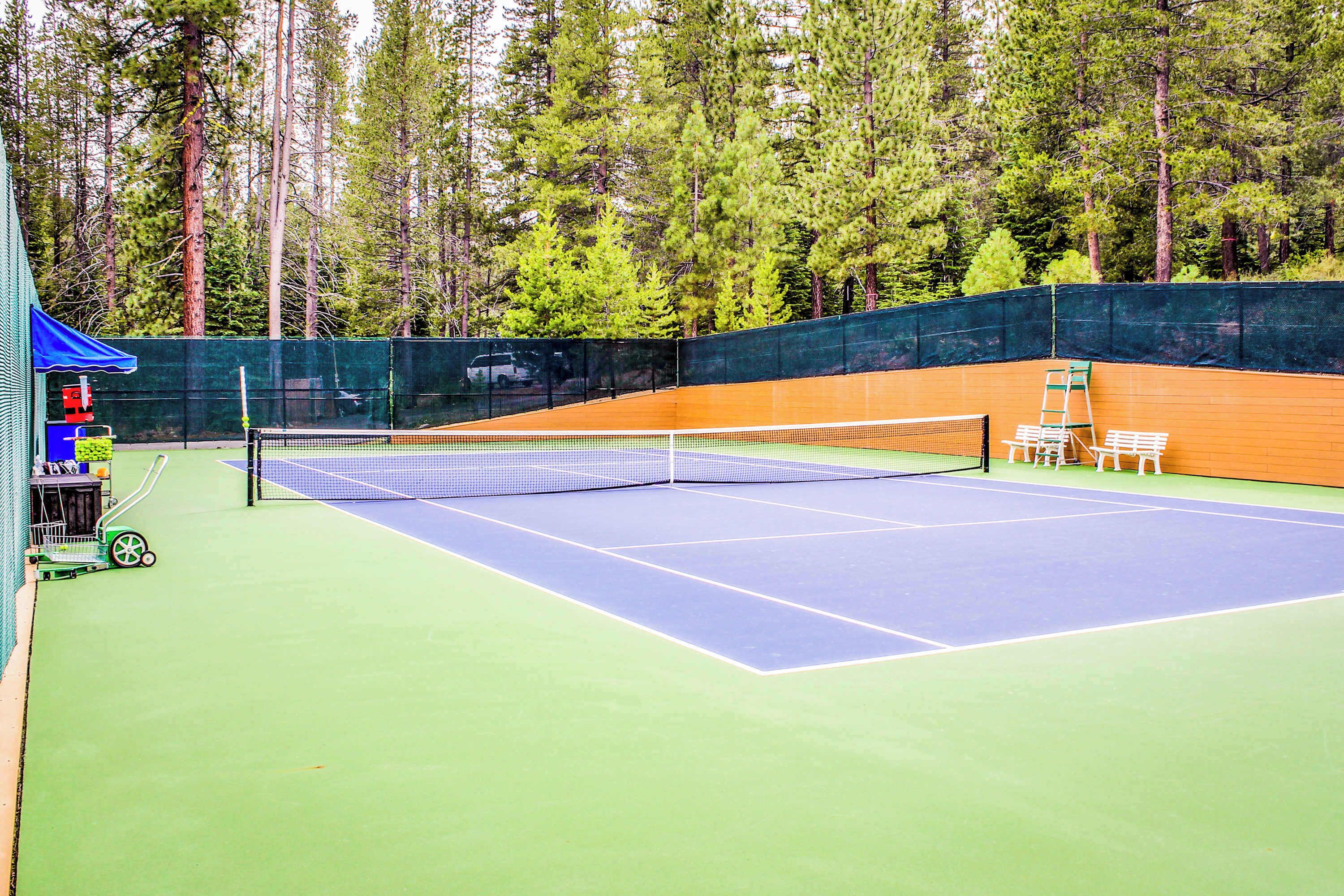 Una cancha de tenis con una red, rodeada por una cerca verde y una tienda de campaña azul al fondo.