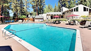 A rectangular swimming pool with a diving board, surrounded by lounge chairs and umbrellas, with a building in the background. - Townhome with pool, sauna, tennis, private hot tub, & foosball, sleeps 8 (Zephyr Cove)