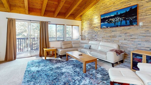 A spacious living room with a wooden ceiling, a stone wall with a painting, a beige sofa, a coffee table, and a view through large windows. - Townhome with pool, sauna, tennis, private hot tub, & foosball, sleeps 8 (Zephyr Cove)