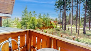 Una terraza de madera con barandilla metálica, que da vista a un área boscosa con árboles altos y una vista distante de casas.