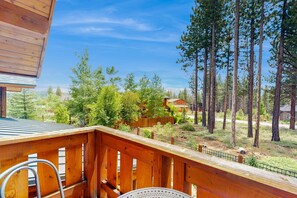 Eine Holzterrasse mit Metallgeländer, die auf einen bewaldeten Bereich mit hohen Bäumen und eine entfernte Ansicht von Häusern blickt.