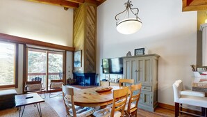 A dining area with a wooden table, chairs, a chandelier, and a view through large windows. - Sophisticated Gold Bend Getaway | Fireplace | Free Ski Shuttle, Pool, Hot Tub (Northstar - Truckee)