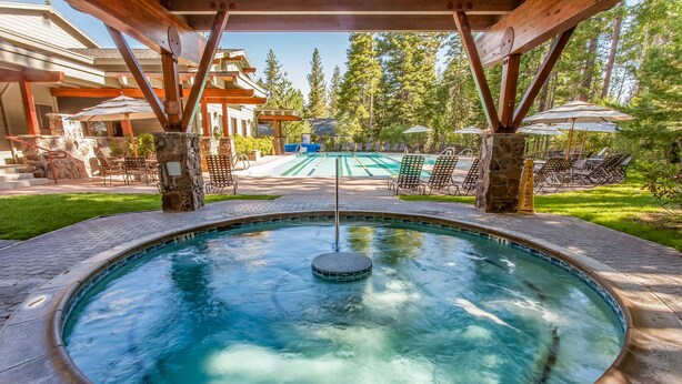 A hot tub with a stone surround, a pool, lounge chairs, and umbrellas in a wooded outdoor area. - Sophisticated Gold Bend Getaway | Fireplace | Free Ski Shuttle, Pool, Hot Tub (Northstar - Truckee)