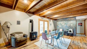 A cozy living room with a fireplace, wooden ceiling beams, and a dining area with chairs and a glass table. - Home with private hot tub, 2 fireplaces, & W&D - drive minutes to hiking, golf (Truckee)
