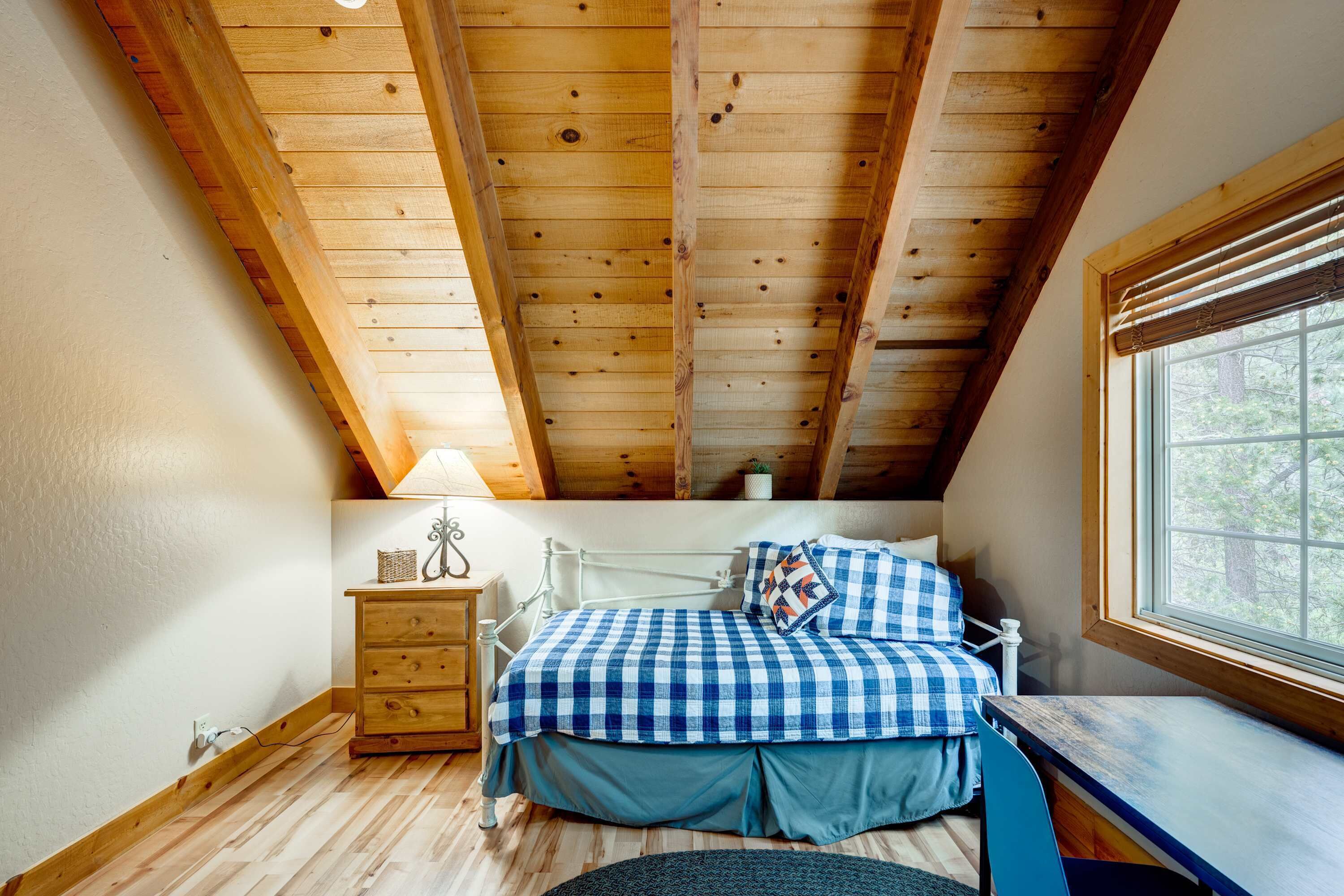 Une chambre avec un plafond en pente, un lit avec une literie à carreaux, une table de chevet, une commode et une fenêtre donnant sur des arbres.