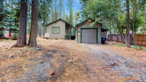 A rustic cabin with a garage, surrounded by tall trees and a gravel driveway. - Tahoe Getaway: Spacious Deck, Fireplace & Family-Friendly (South Lake Tahoe)