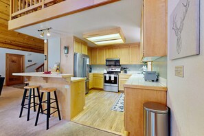 A compact kitchen with wooden cabinets, a stainless steel refrigerator, a microwave, and a stove. There are two stools at the counter and a trash can to the side. - Cozy family-friendly home with wood-burning fireplace, loft, desk, & pool access (Truckee)