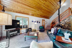 A living room with a wooden ceiling, a television, a stone fireplace, a leather sofa, and a staircase. - Cozy family-friendly home with wood-burning fireplace, loft, desk, & pool access (Truckee)