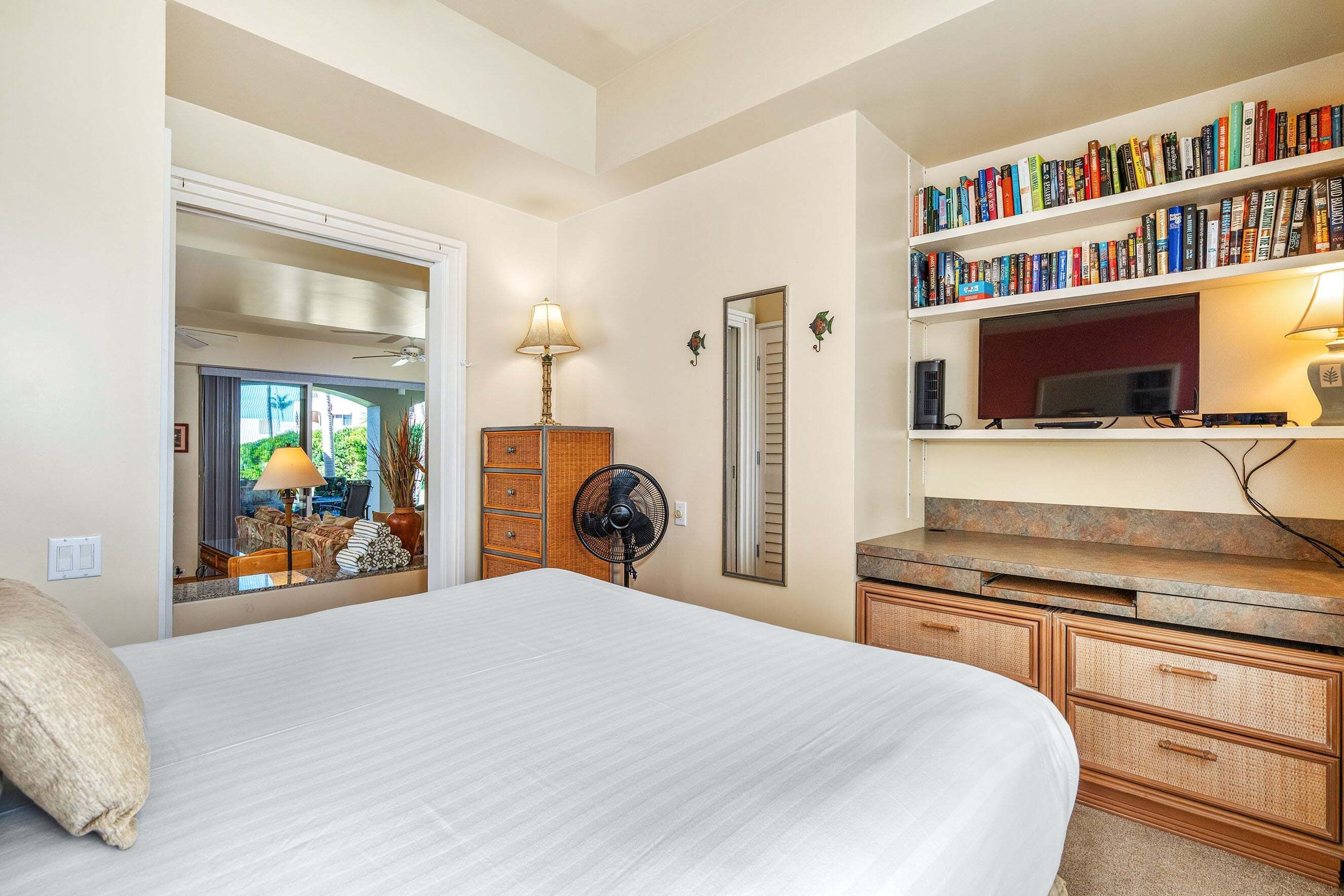 A hotel room with a bed, a bookshelf, a desk, and a fan.