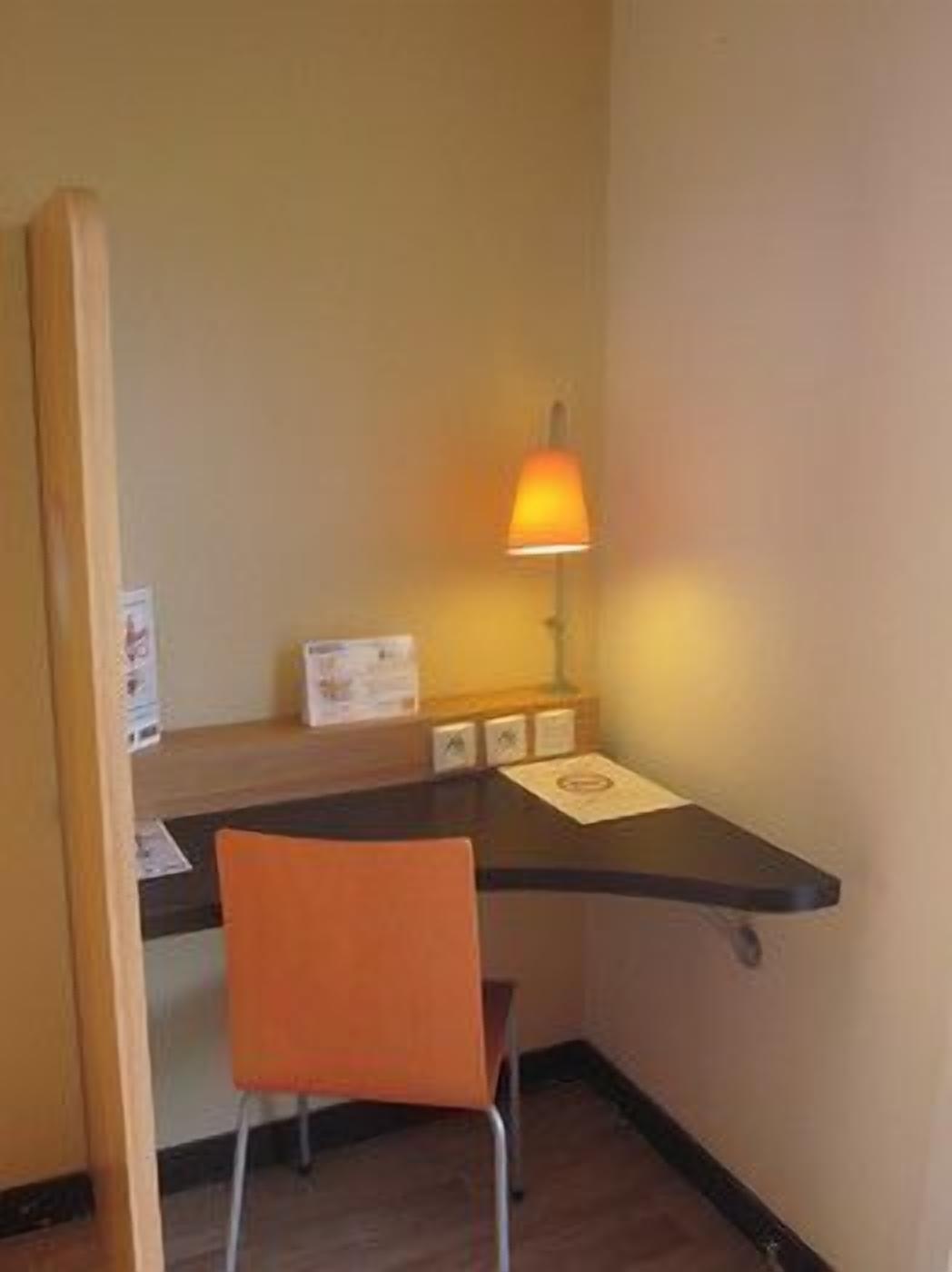 A small hotel room with a desk, chair, and a wall-mounted lamp.