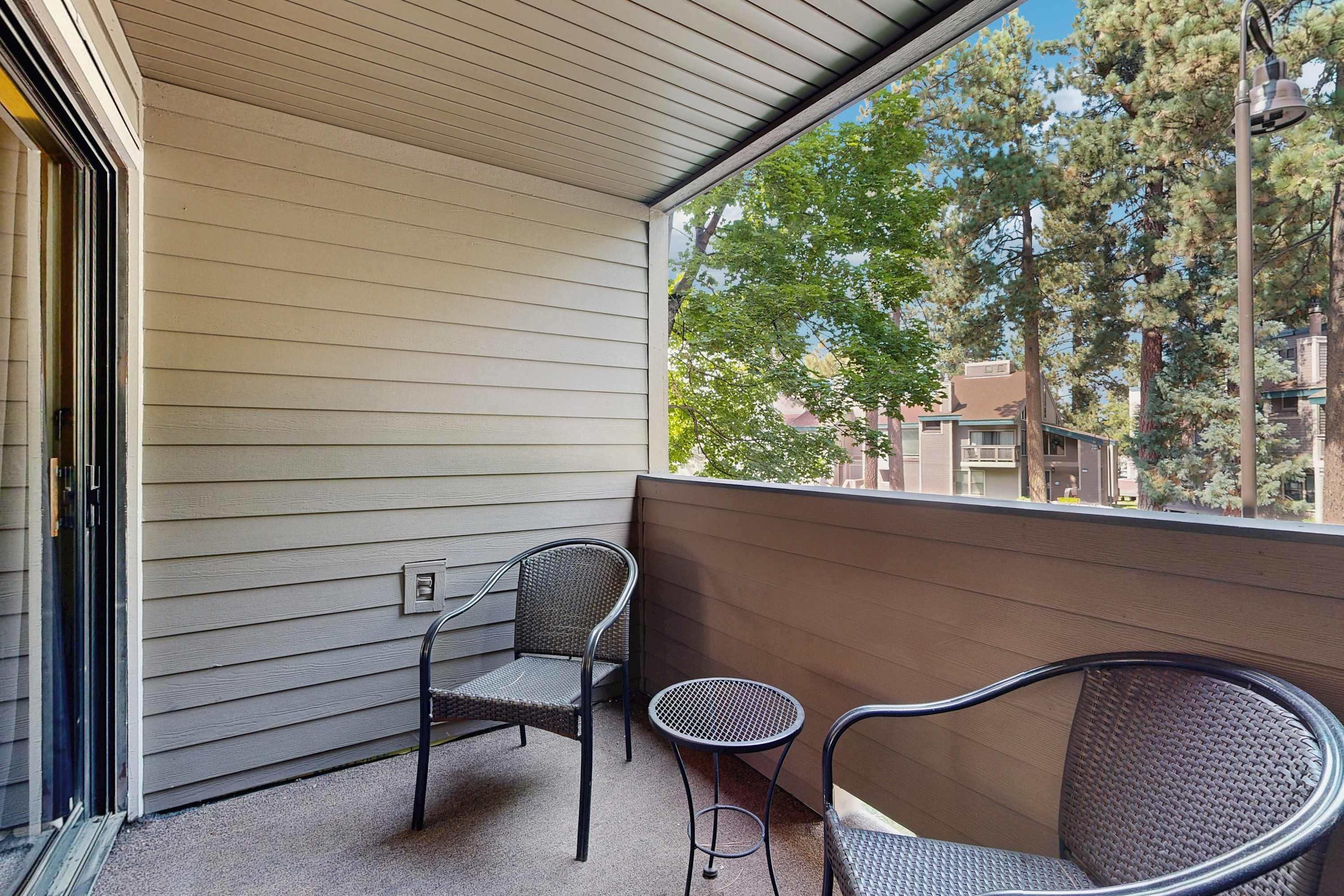 Ein Balkon mit zwei Stühlen und einem kleinen Tisch, ein Fenster mit Blick auf Bäume und Gebäude sowie eine Tür mit Glasfenster.