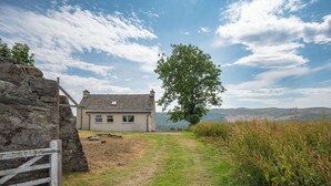En stenhytte med en græsplæne, der fører hen til den, omgivet af vegetation og en klar himmel.