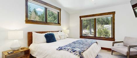 Un dormitorio con cama, dos ventanas, ventilador de techo, mesita de noche y una silla.