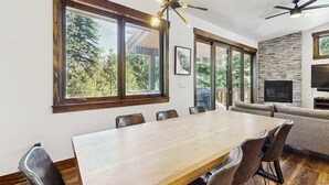 Ein Essbereich mit einem großen Holztisch, mehreren Stühlen, einem Deckenventilator und großen Fenstern mit Blick auf Bäume.