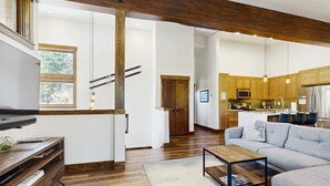Ein modernes Wohnzimmer mit einer Couch, einem Couchtisch und Holzböden. Im Hintergrund ist eine Küche mit Holzschränken und Geräten zu sehen.