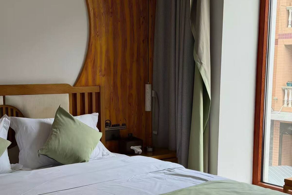 Una habitación de hotel con una cama grande, cabecero de madera, una mesita de noche, una lámpara y una ventana con vistas a edificios.