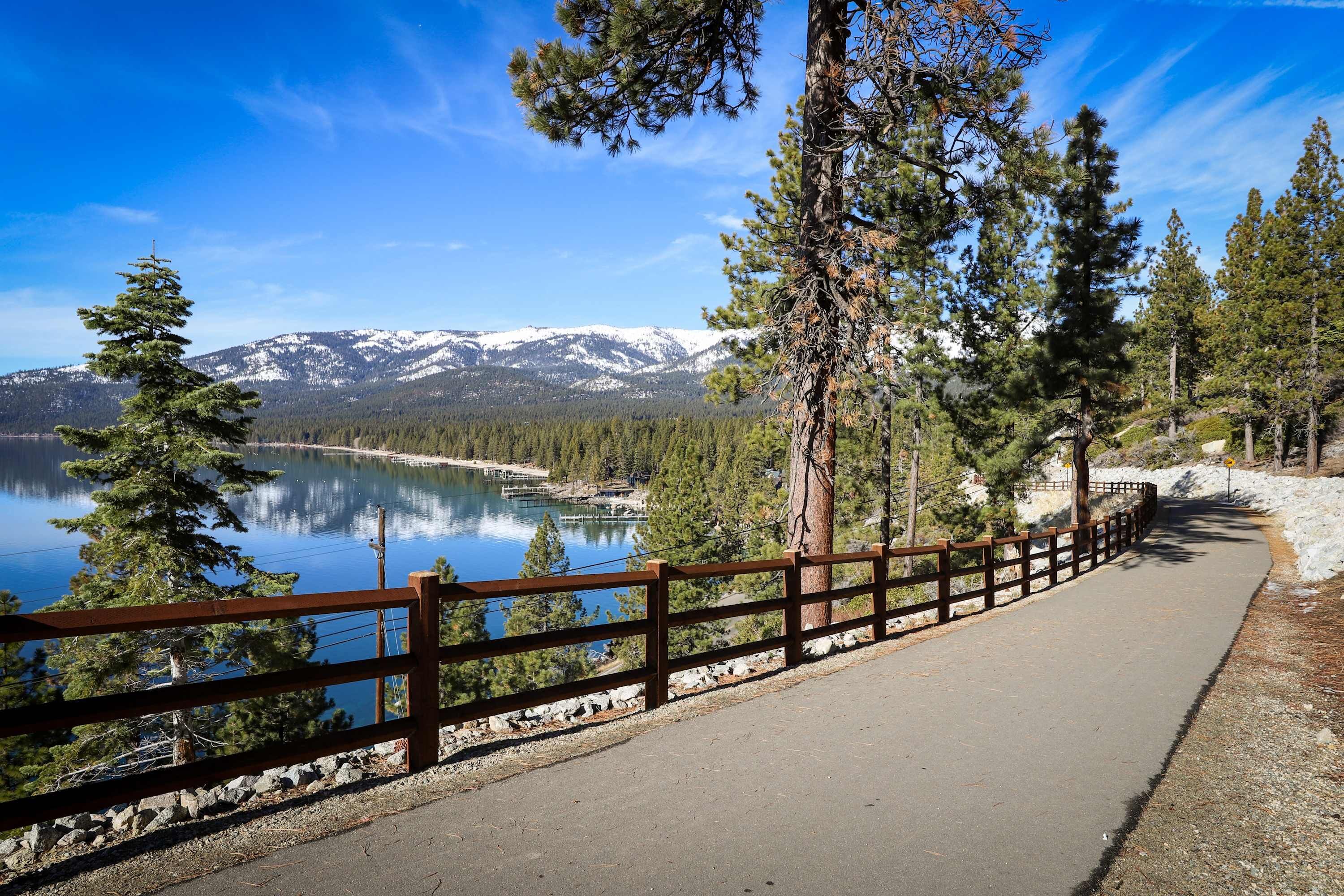 A paved walkway with a wooden railing, surrounded by trees and a lake with mountains in the background.