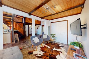Uma sala de estar ampla com teto de madeira, tapete de couro, duas poltronas cinzas, uma televisão e uma planta.