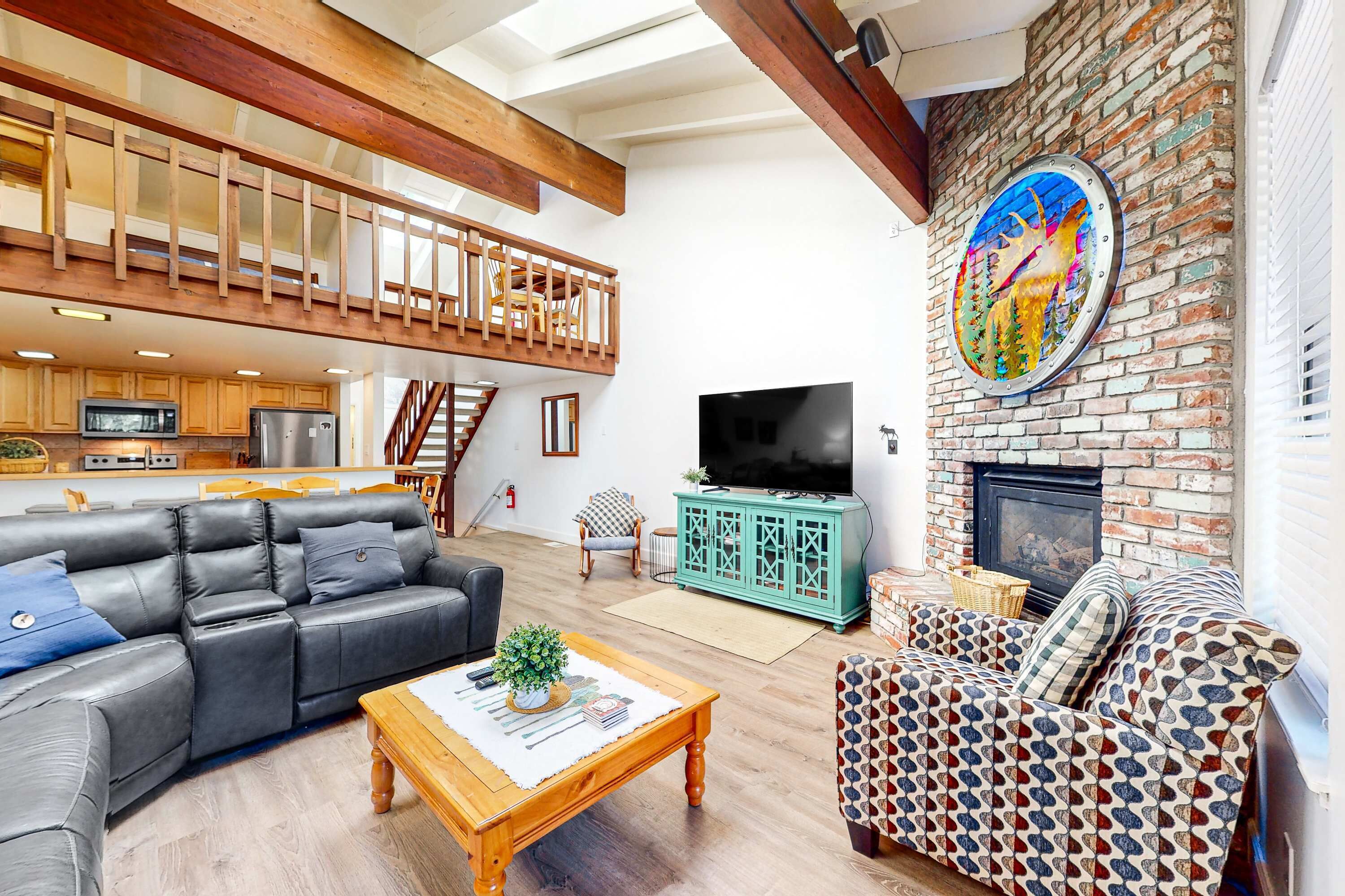 A spacious living area with a black leather sofa, patterned armchair, and a wooden coffee table. A television and fireplace are visible, along with a kitchen in the background.
