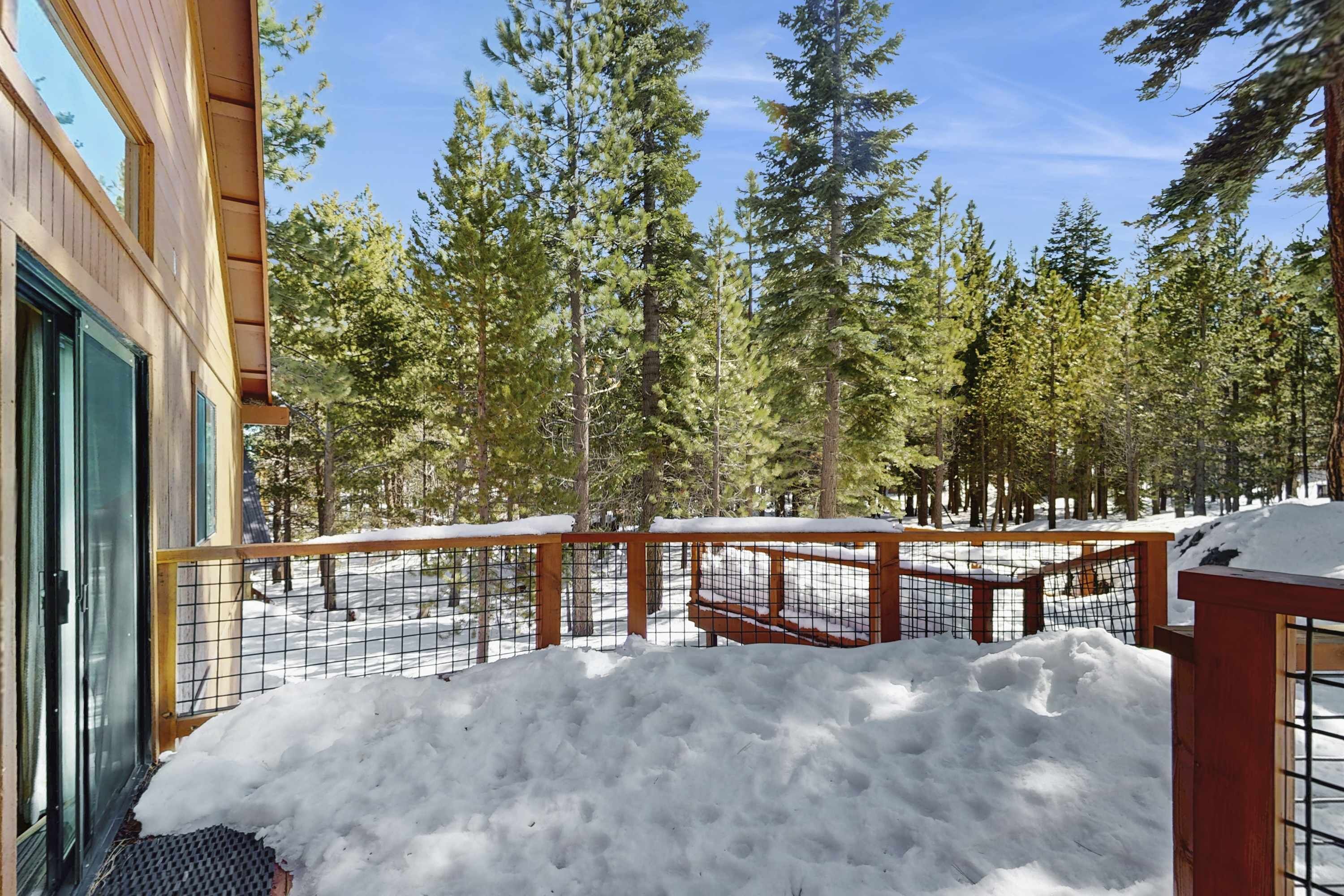 Una terraza de madera con baranda de metal, rodeada de nieve y árboles.