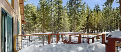 Una terraza de madera con baranda de metal, rodeada de nieve y árboles.