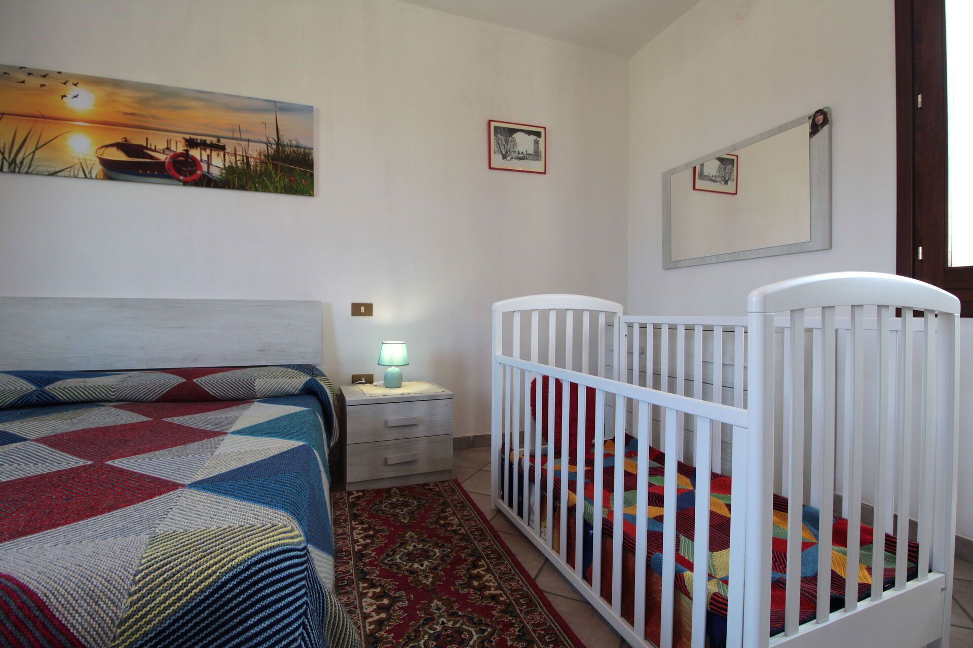 A bedroom with a bed, a crib, a nightstand, a lamp, a painting, and framed pictures on the wall.